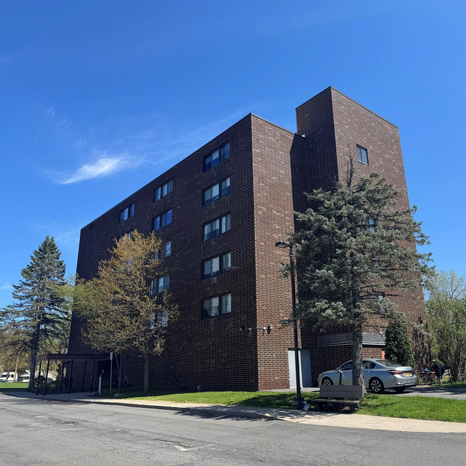 Brown brick apartment building with trees and parked cars on a sunny street