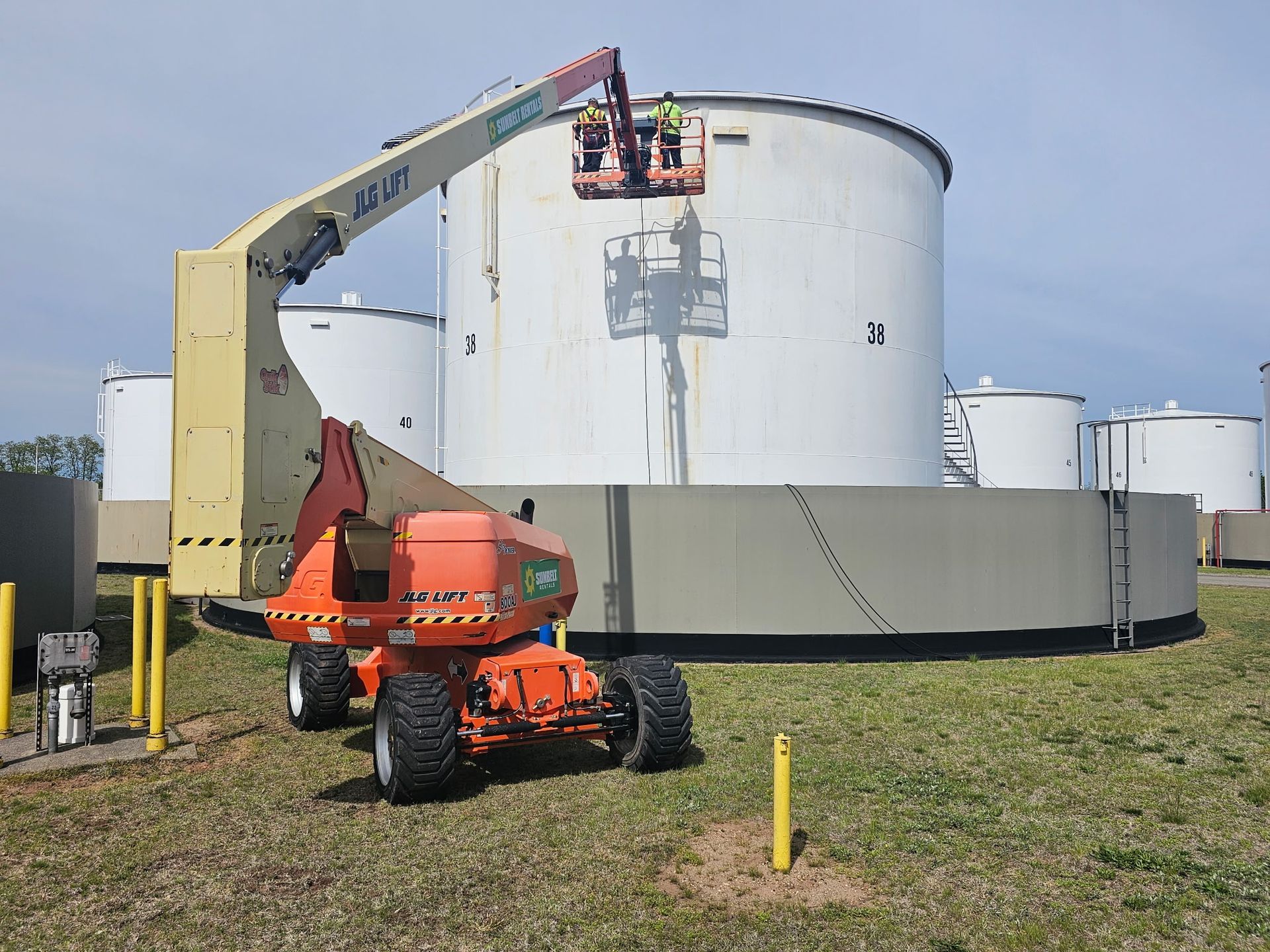 Orange boom lift servicing a large white storage tank in an industrial field.