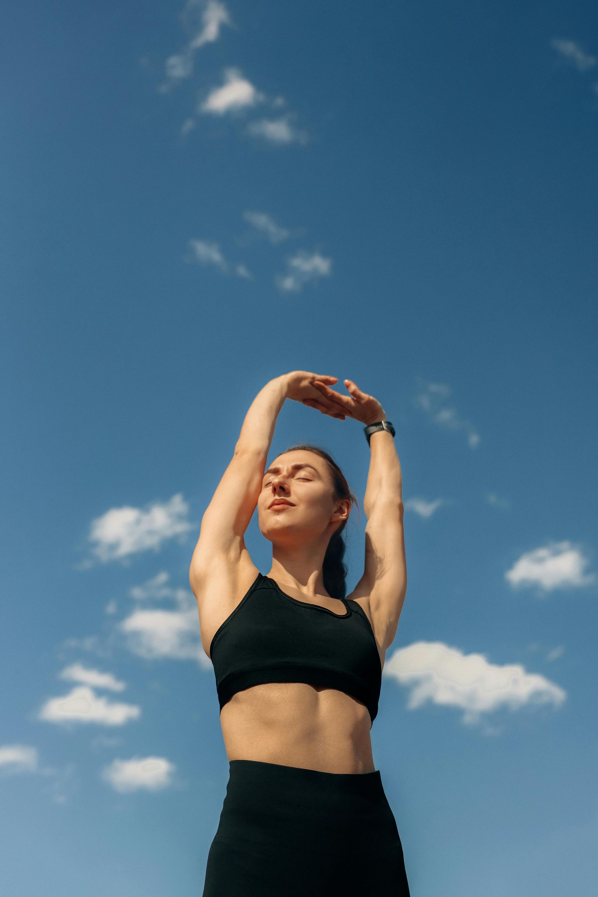 Person stretching outdoors against a blue sky with clouds, wearing a black sports top.