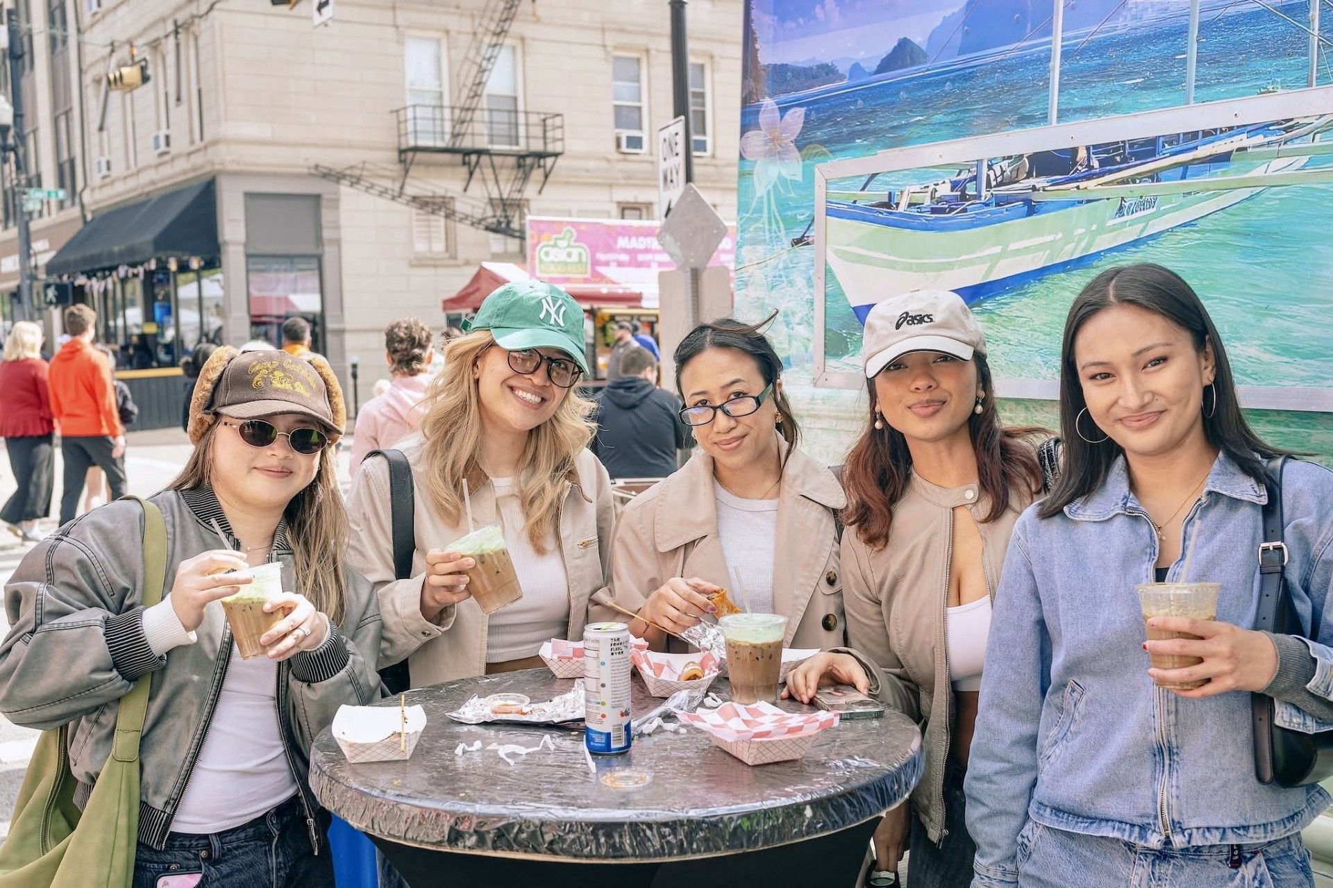 Five friends smiling at a café table outdoors, with drinks and a waterfront mural in the background.