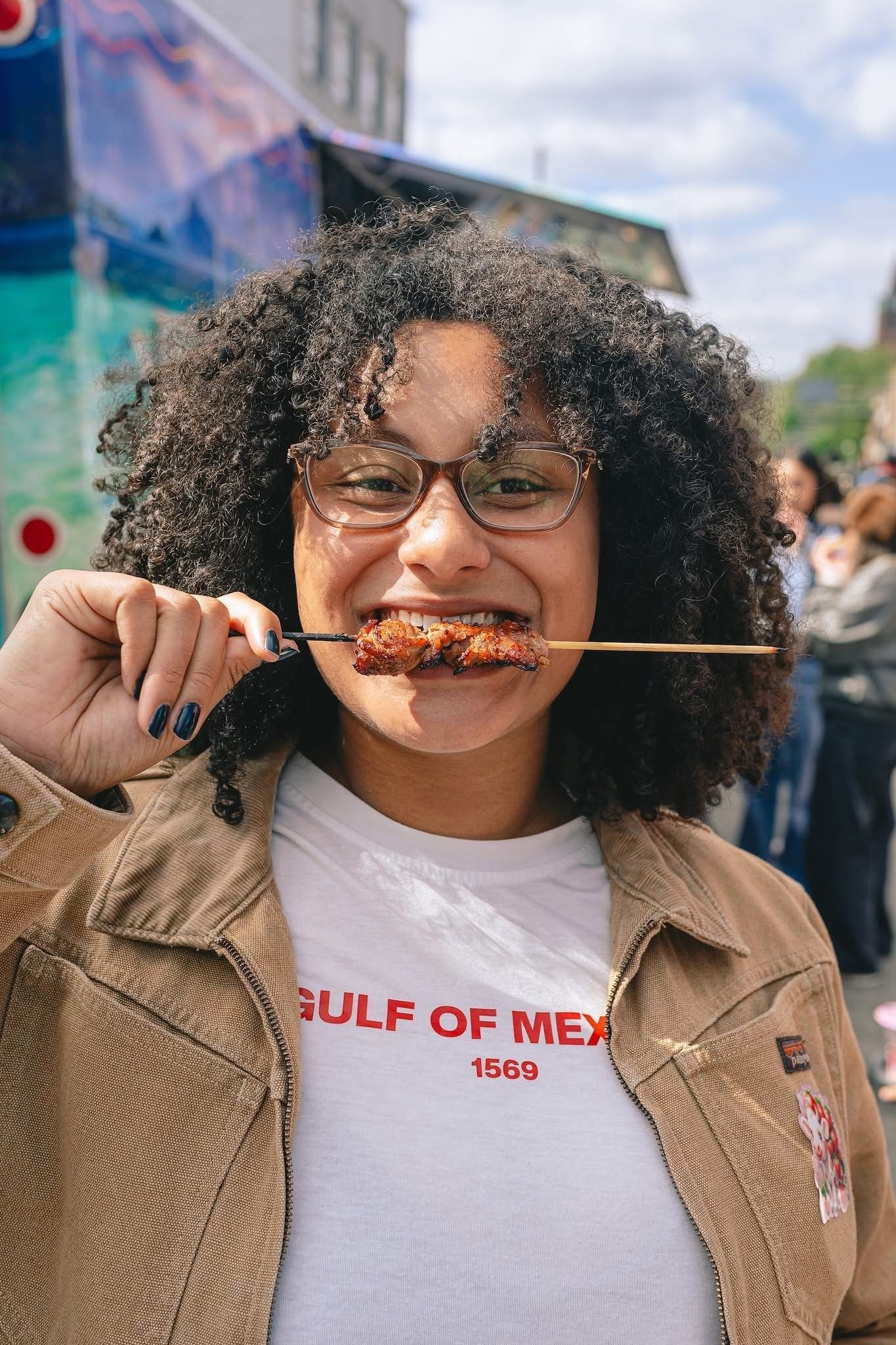 Person smiling while holding a skewer of food at an outdoor event, wearing glasses and a brown jacket.