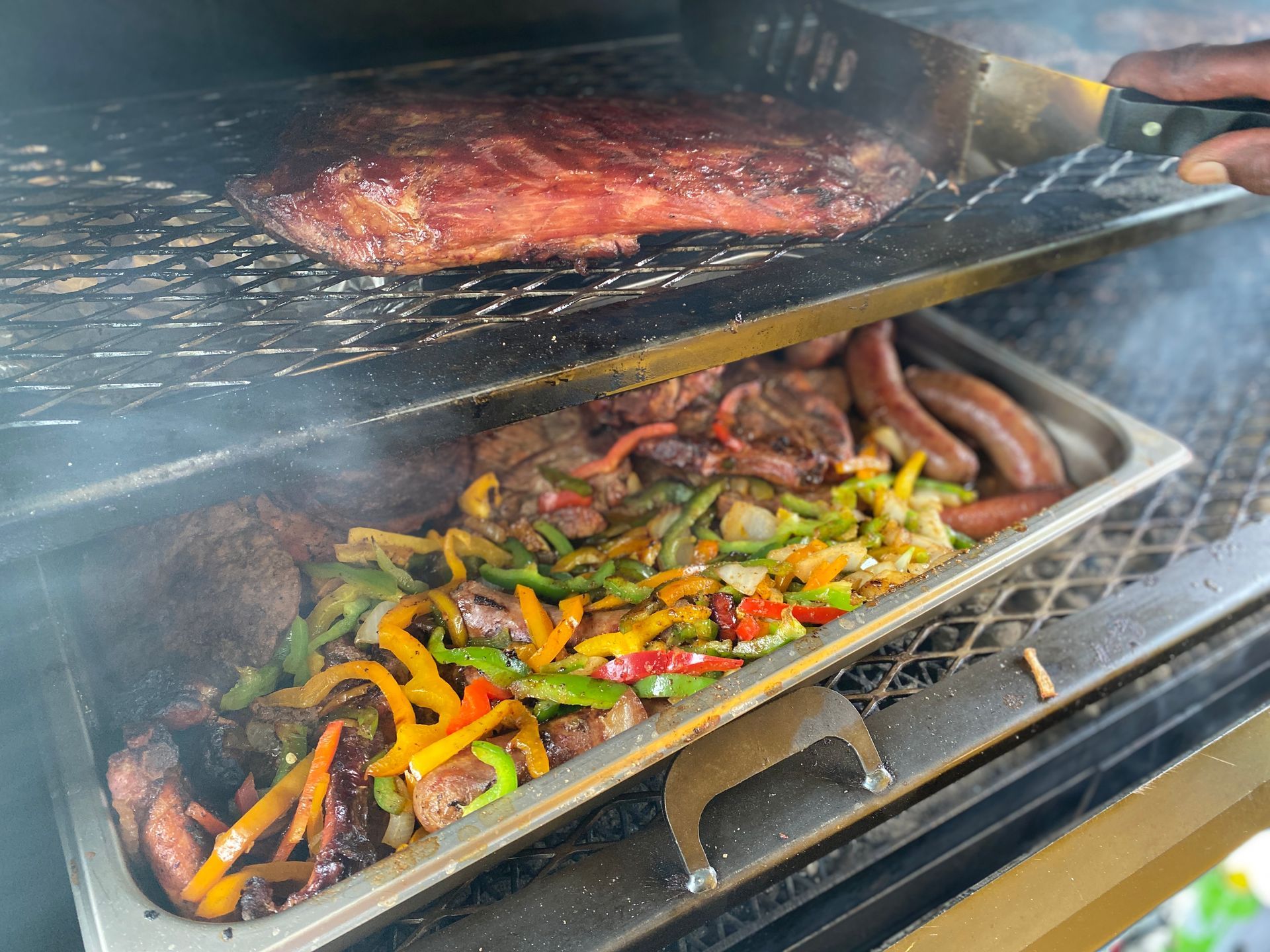 Open grill with sizzling steak and sausages beside colorful sautéed peppers and onions