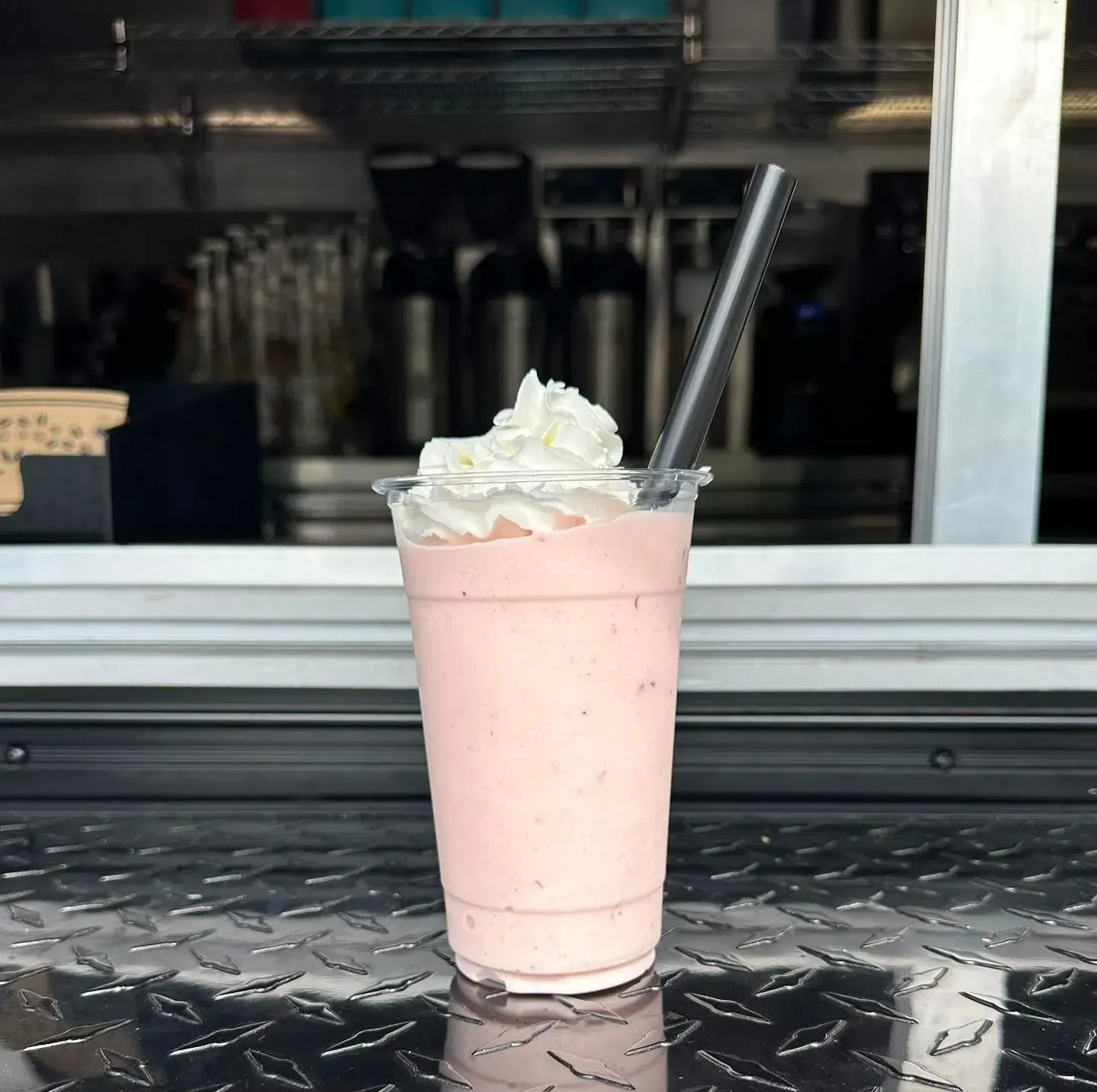 Pink milkshake in a clear cup with whipped cream and a striped straw on a metal counter.