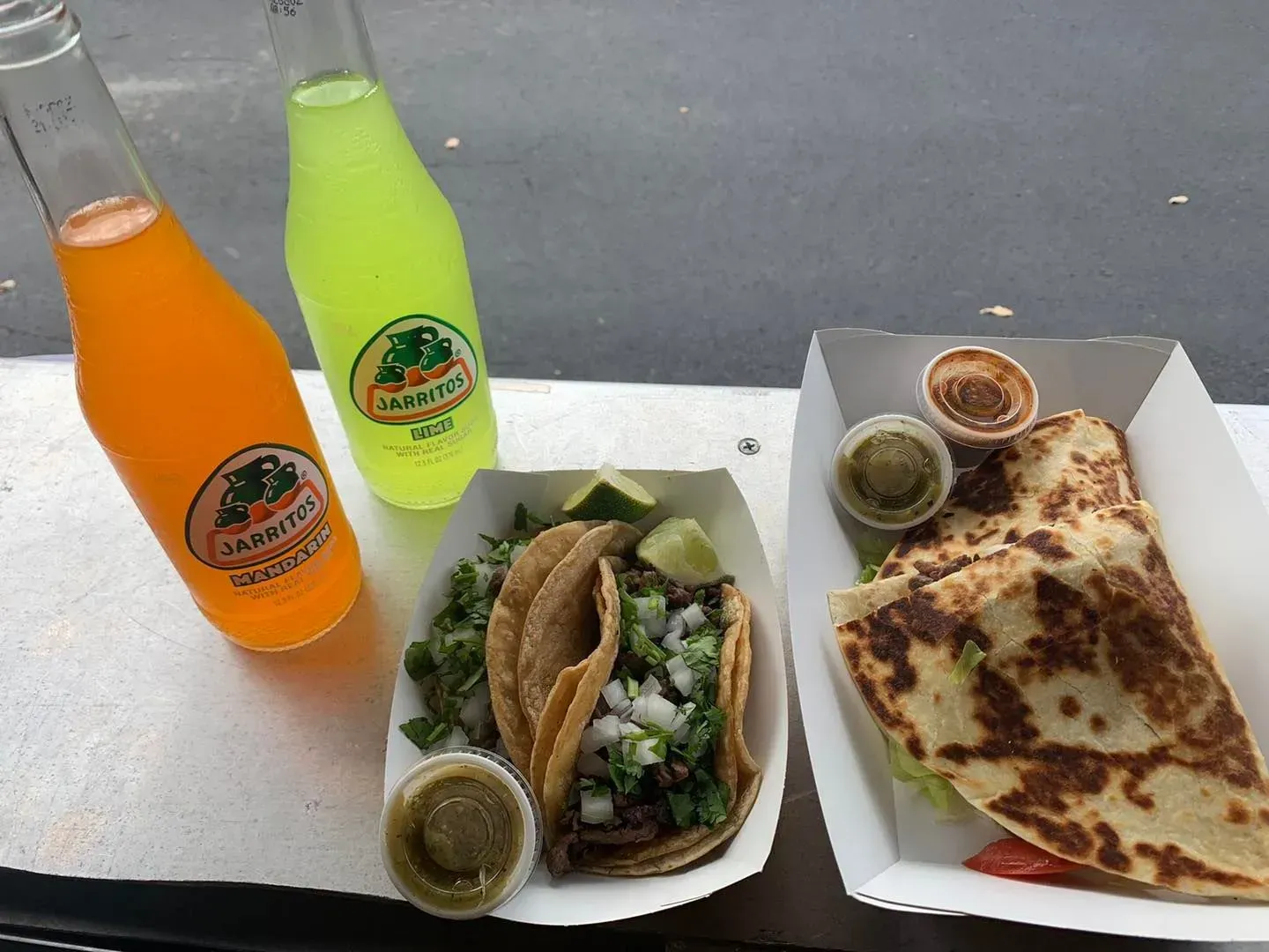 Two bottled drinks and takeout food on a table, including tacos, quesadilla, and salsa.