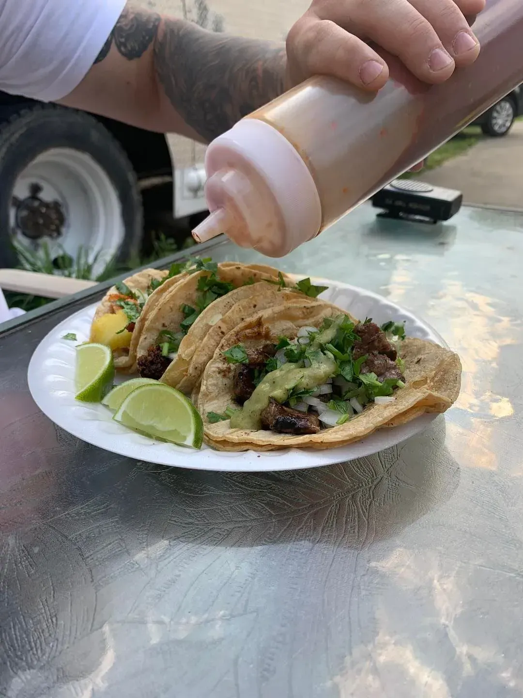 Tacos on a plate being topped with sauce at an outdoor table with lime wedges