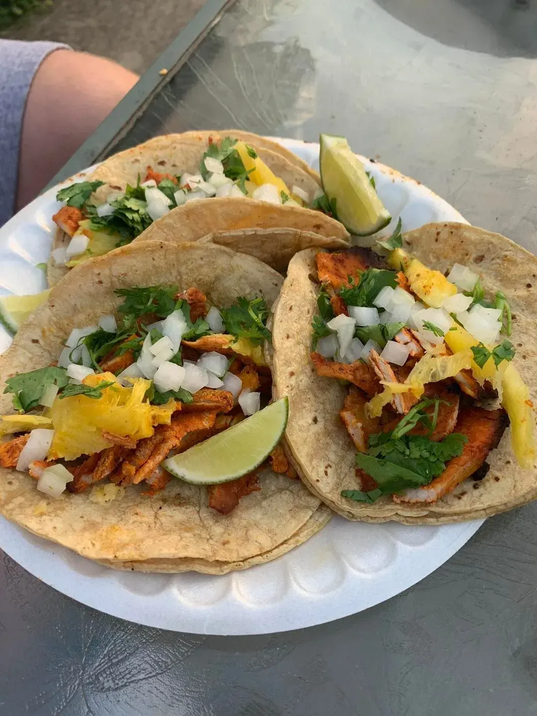Three tacos on a white plate with lime wedges and chopped onions, cilantro, and salsa on a table