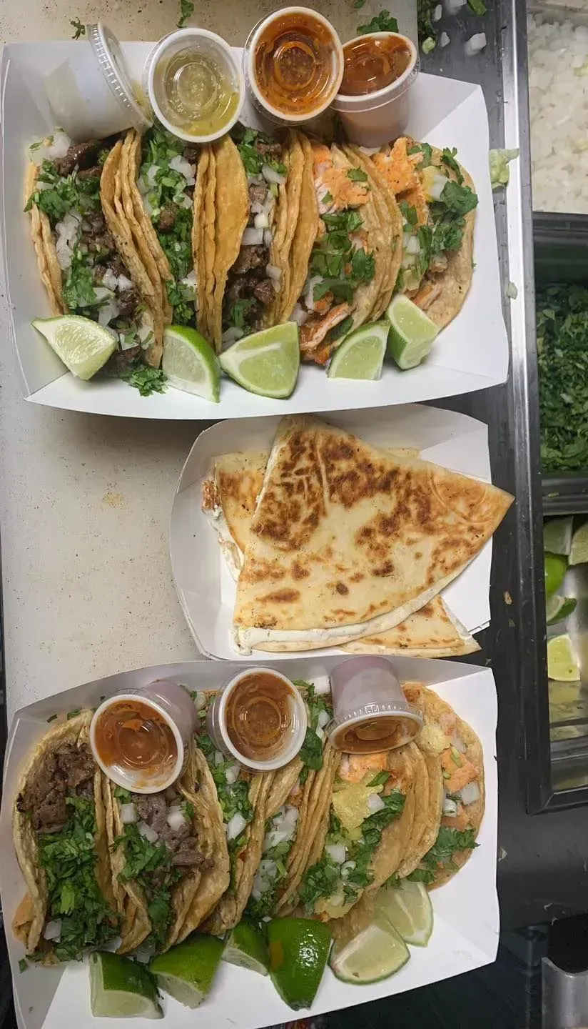 Three plates of tacos with limes and sauces, plus a quesadilla on a table near a window.