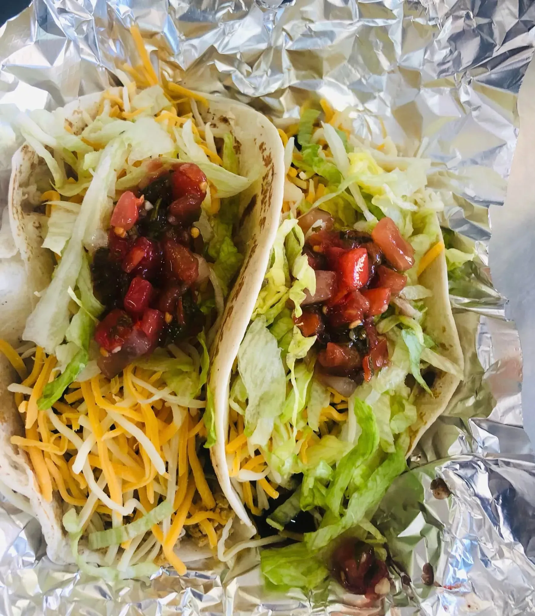 Two tacos in foil, topped with lettuce, shredded cheese, and red salsa
