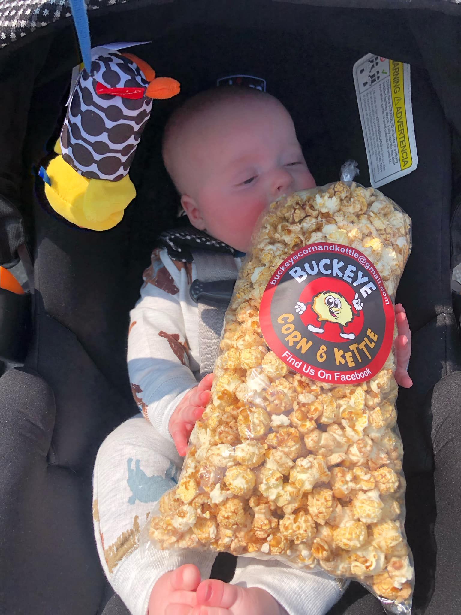 Baby in a car seat holding a bag of Bucc-ee's caramel popcorn with a giraffe toy nearby