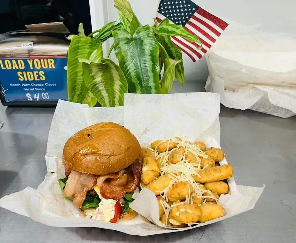 Burger and loaded fries in a takeout basket on a counter, with an American flag and plant in the background