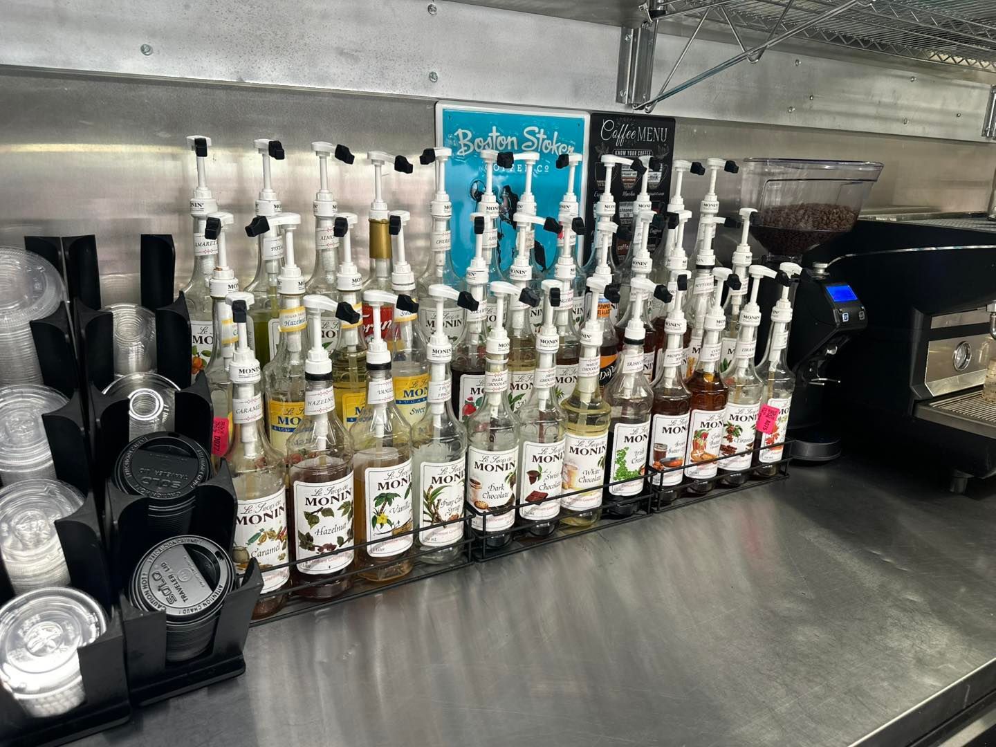 Coffee shop counter lined with syrup bottles, cups, and a drink machine behind a stainless-steel counter