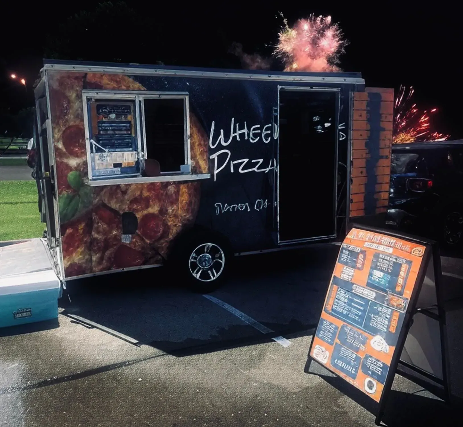 Mobile pizza food truck with lit menu board at night, fireworks bursting overhead.