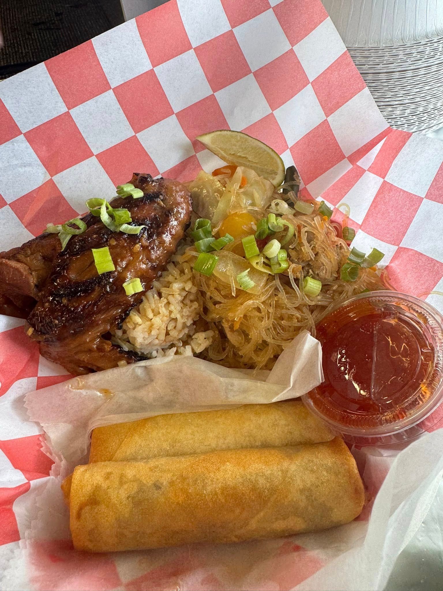 Plate of glazed grilled meat with rice, sauce, and two spring rolls on red-and-white checkered paper