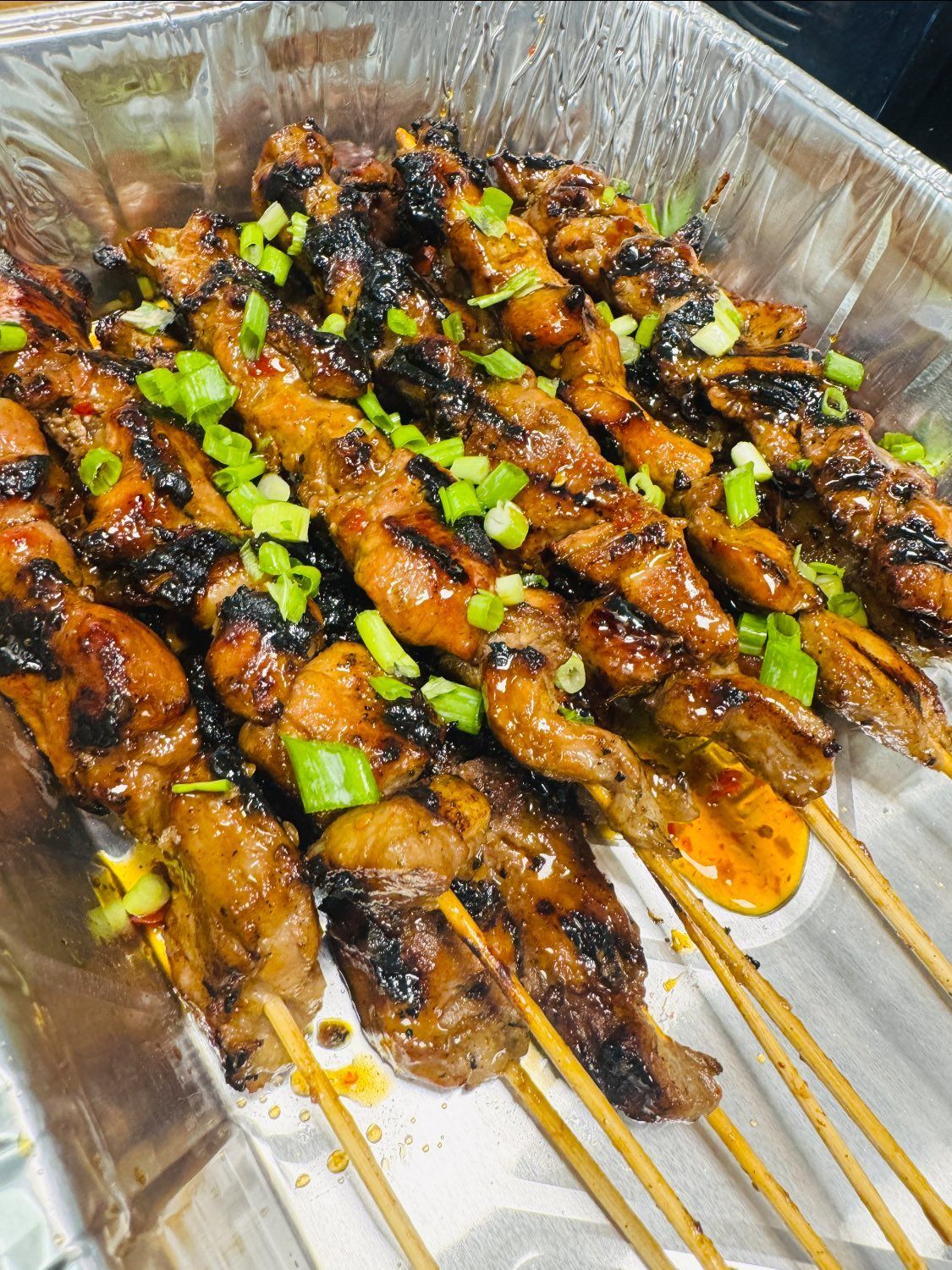 Skewered glazed chicken satay topped with chopped green onions in a foil tray