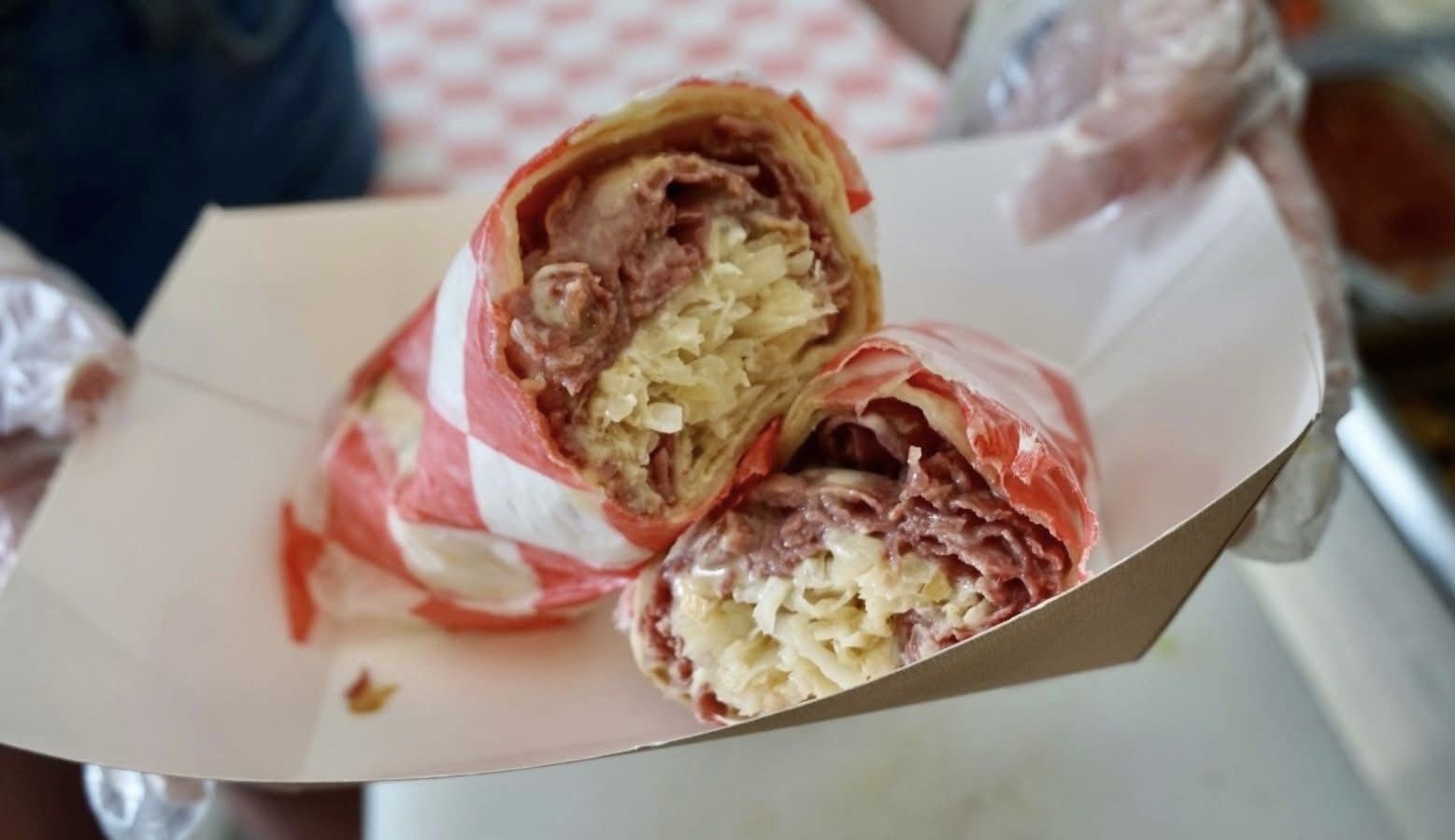 Open sandwich with red checkered paper in a cardboard takeout box, held by gloved hands.
