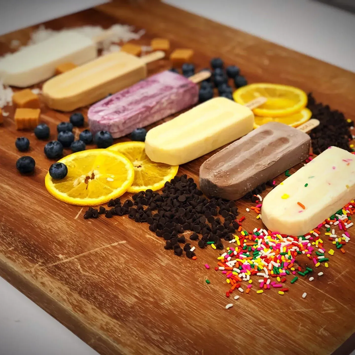 Assorted ice cream bars on a wooden board with orange slices, blueberries, chocolate crumbs, and sprinkles
