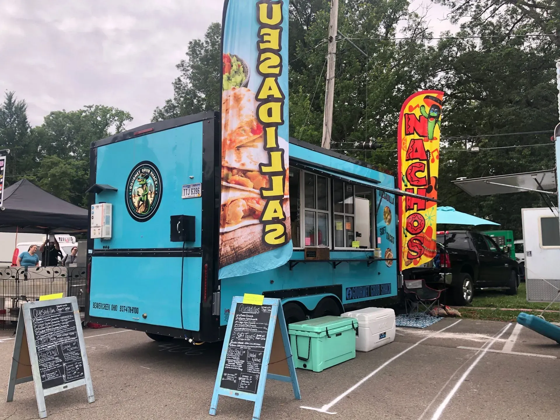 Bright blue food truck with colorful signs at an outdoor market, with chalkboard menus out front