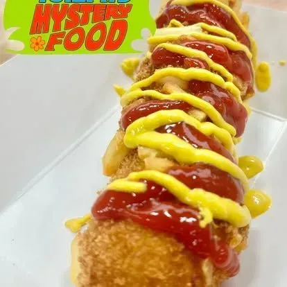 Ketchup and mustard-drizzled corn dog on a white tray, with “Mystery Food” text in the corner