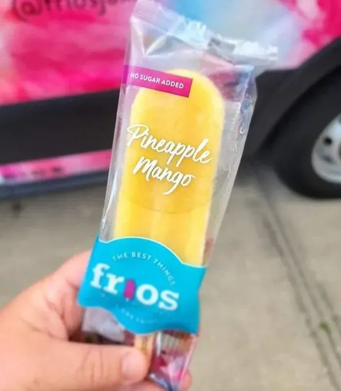 Hand holding a Pineapple Mango frozen treat in clear wrapper with blue Fros label outdoors