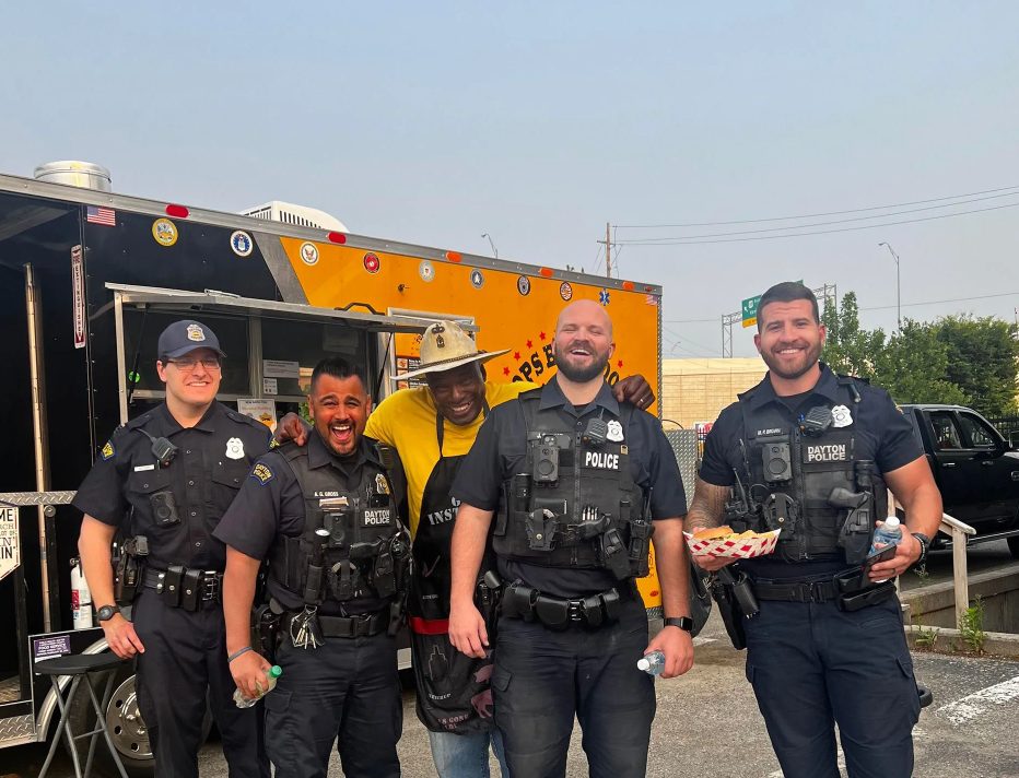 Four police officers in black uniforms stand smiling beside a black and yellow police vehicle.