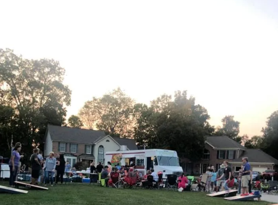 Neighbors gathered on a grassy lawn at dusk for an outdoor community event, with chairs, tents, and a truck stage.