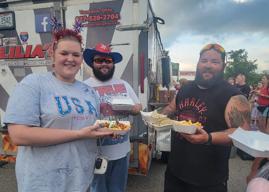 Three people holding plates of food at an outdoor street event, smiling at the camera.
