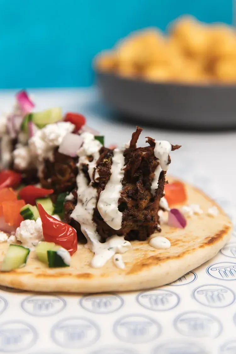 Topped flatbread with meat, vegetables, and sauce on a table, with a bowl of fries blurred in the background