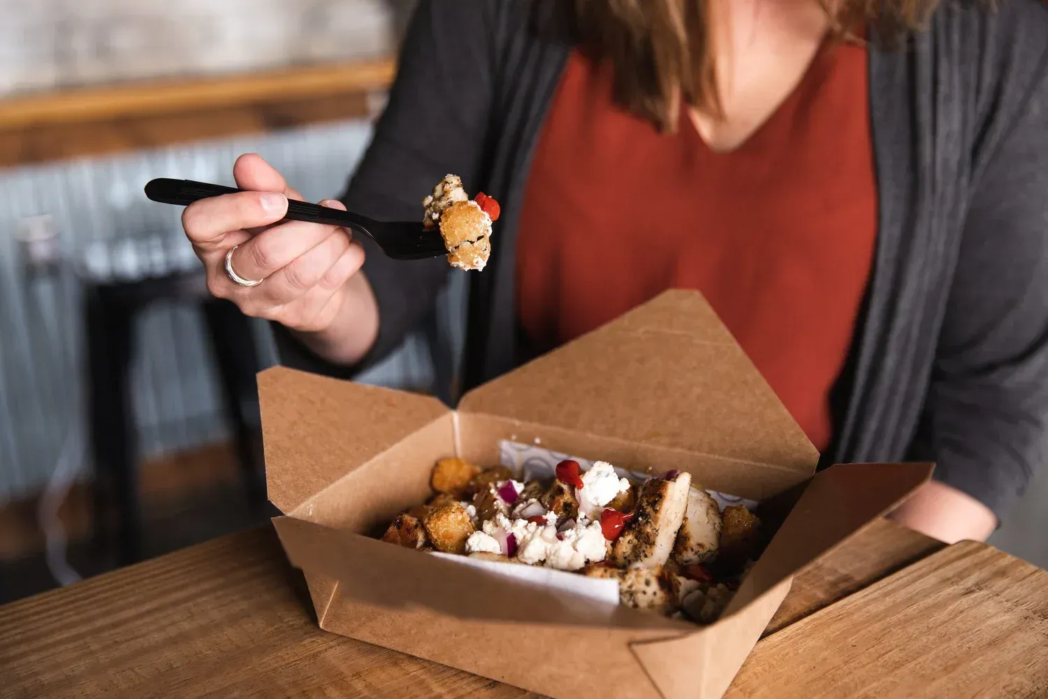 Person eating takeout from a cardboard box with a fork at a wooden table