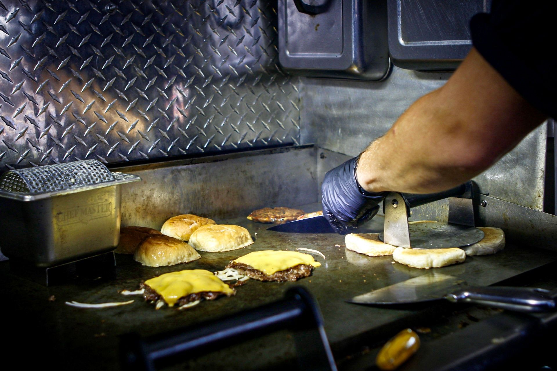 Gloved cook turning burgers and buns on a flat-top grill in a food truck kitchen