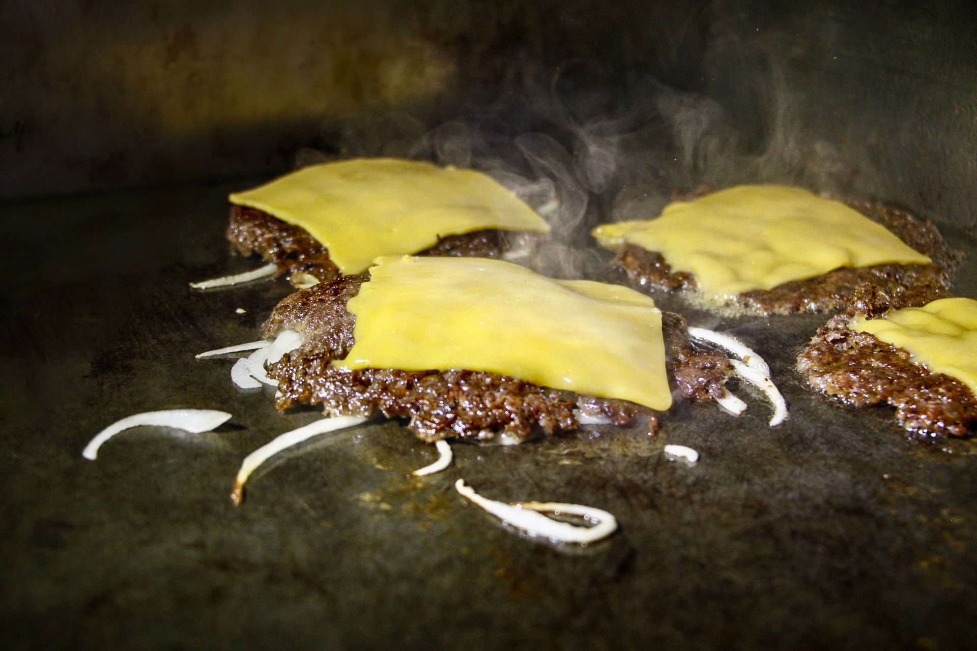 Three cheeseburgers with melted yellow cheese on a dark griddle, with sizzling onions around them