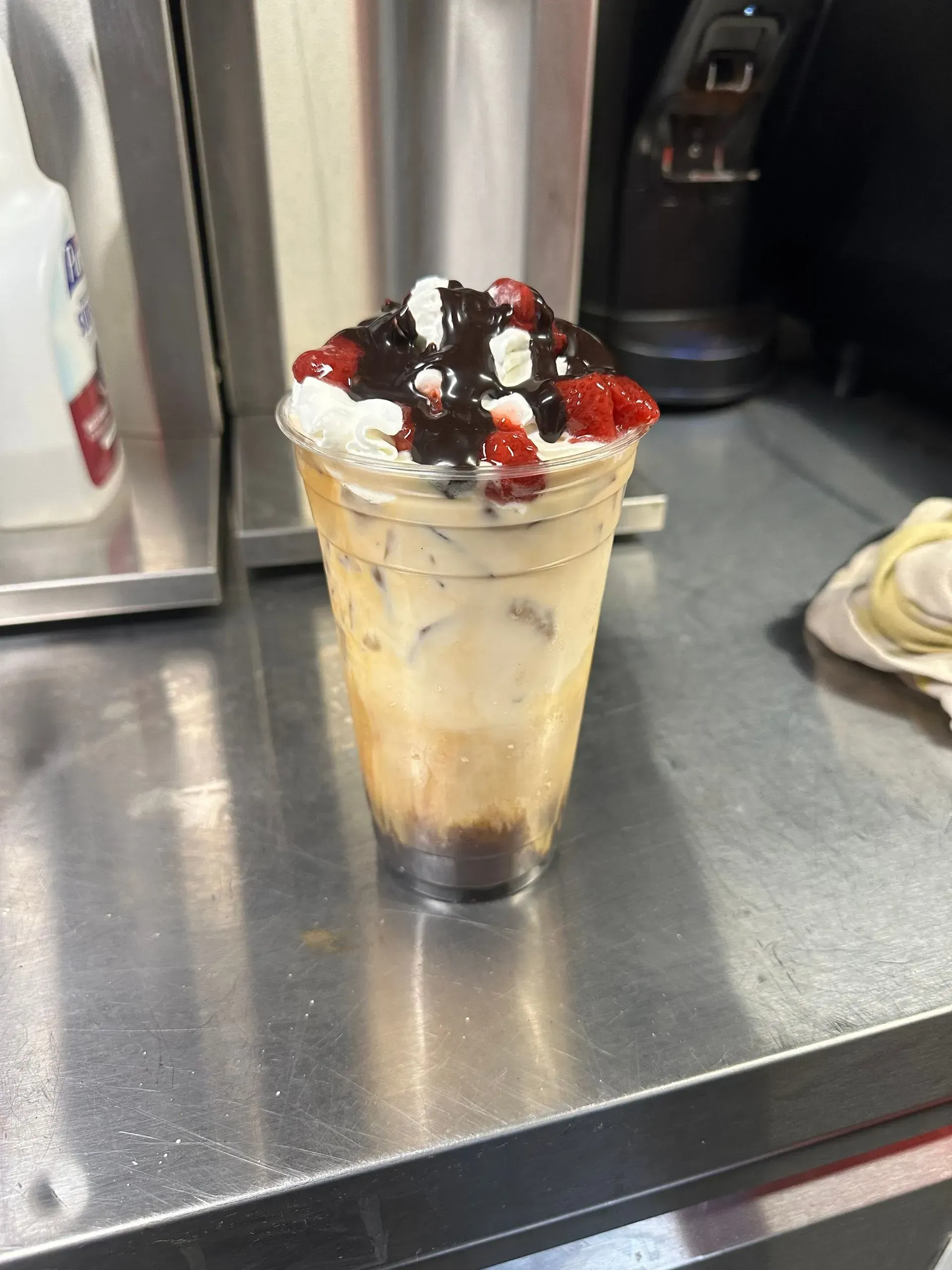 Tall dessert drink topped with whipped cream, dark berries, and red sauce on a metal counter