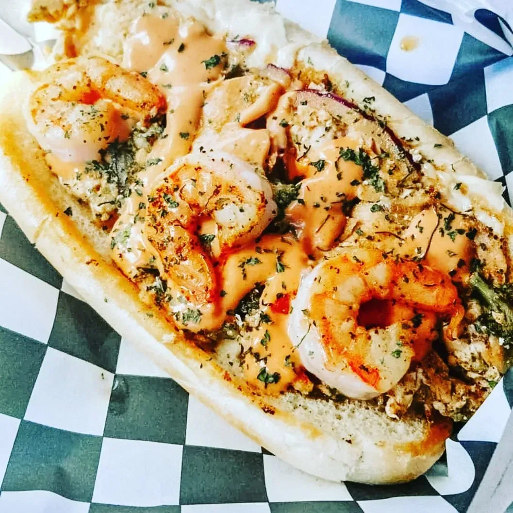 Grilled shrimp po’ boy sandwich on checkered paper, topped with herbs and sauce