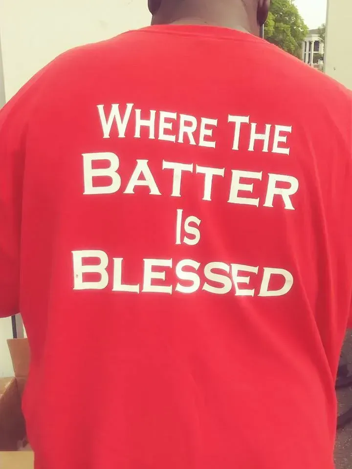 Red T-shirt with white text: “WHERE THE BATTER IS BLESSED”