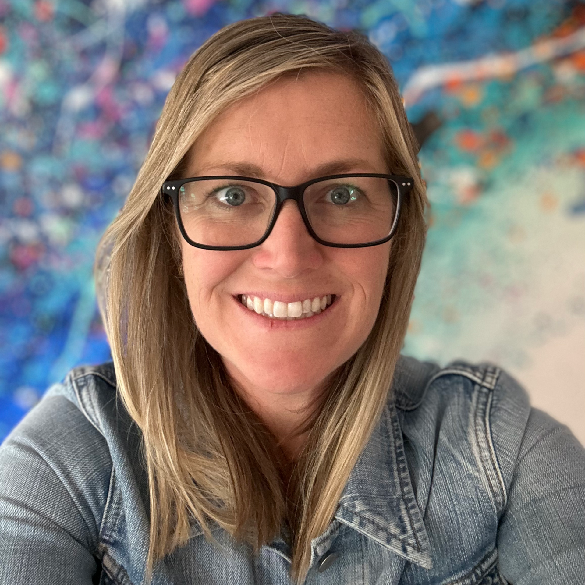 Woman with blonde hair and glasses smiles, wearing a denim jacket; abstract blue and colorful background.