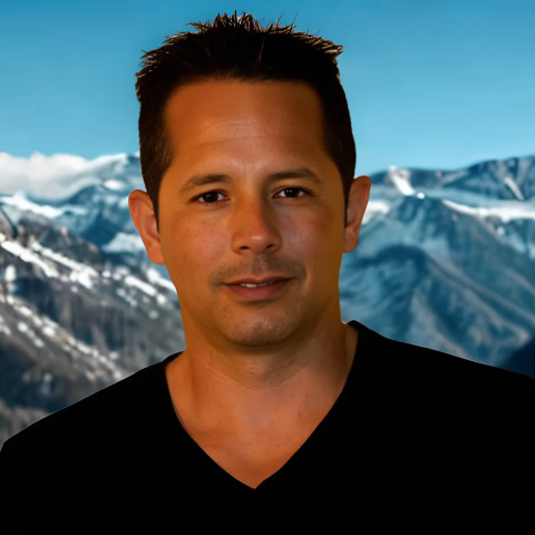 Man with dark hair, smiling, in front of a mountain range. He wears a black V-neck shirt.