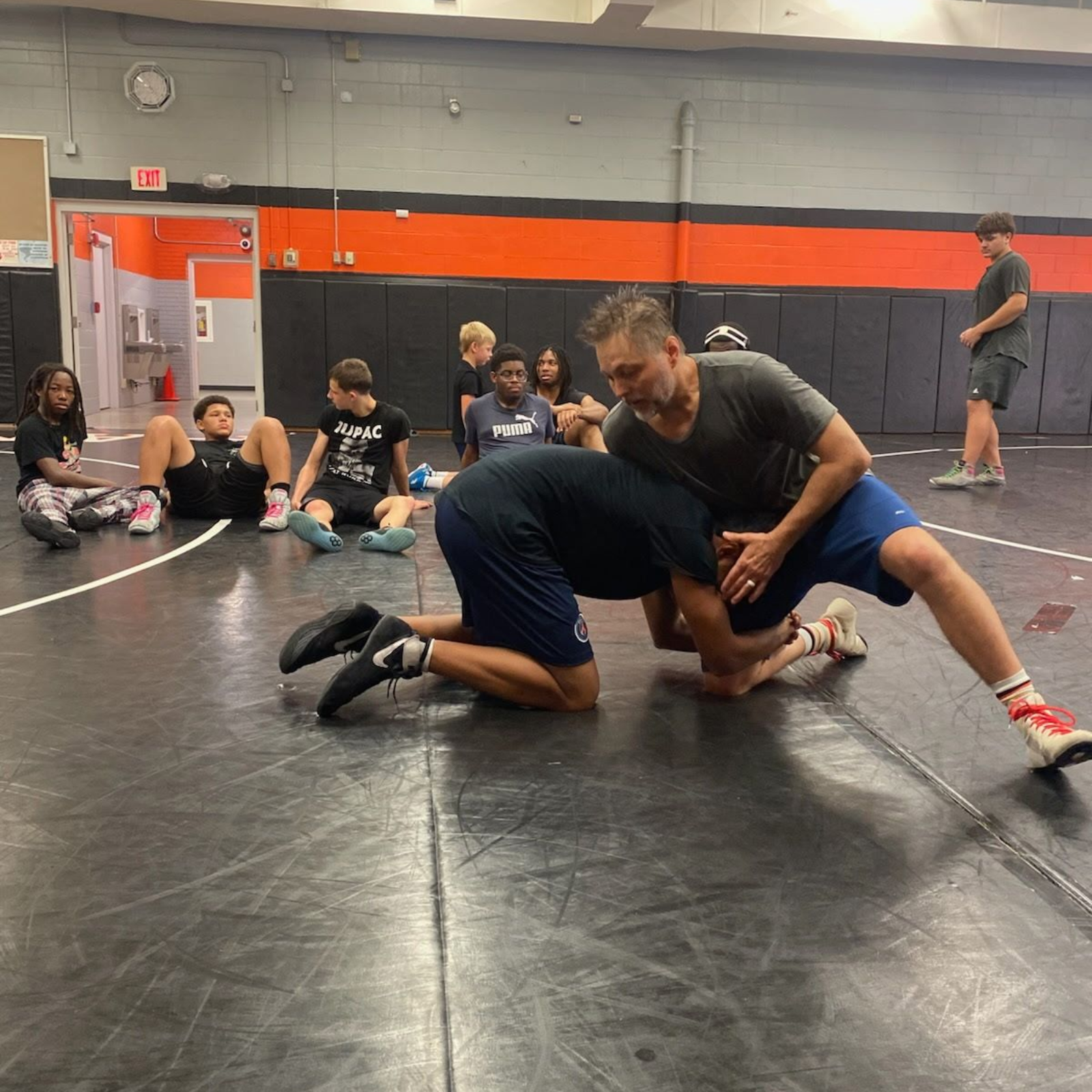 Two men wrestling on a mat in a gym. Other people watch.