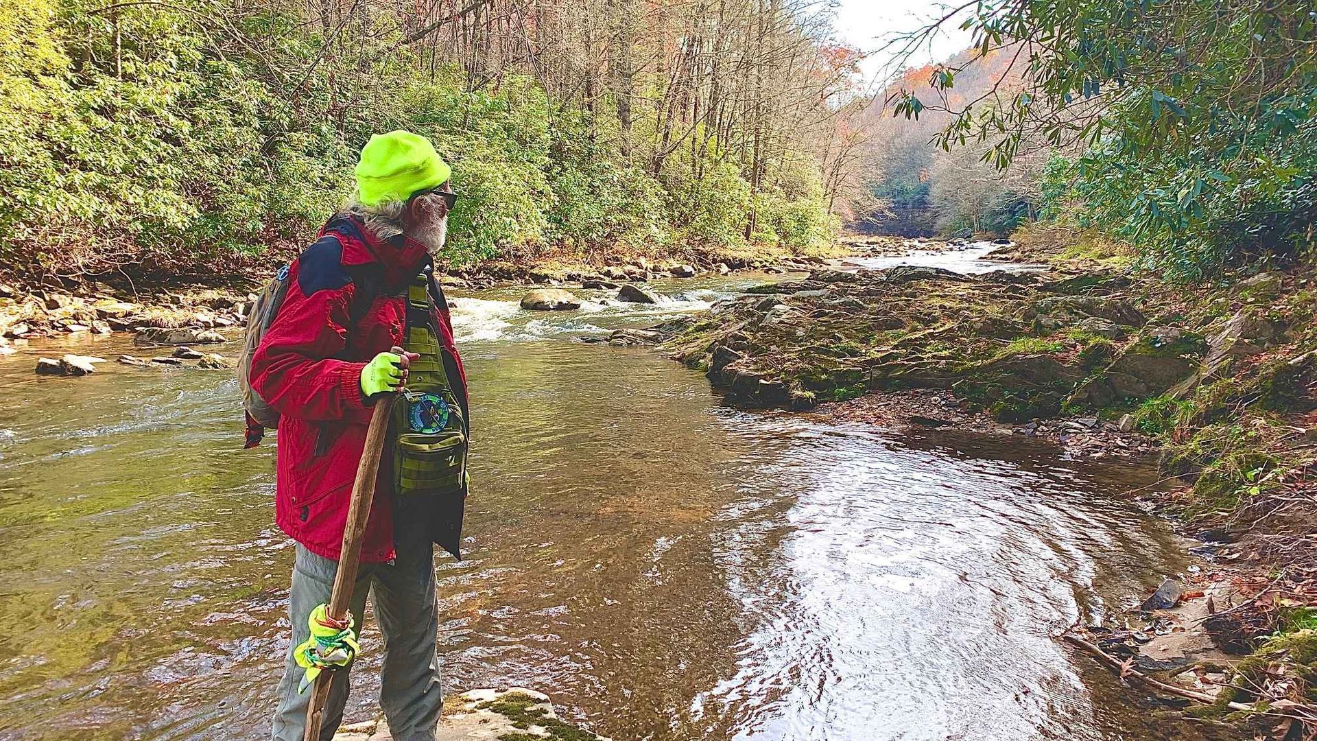 A man in a red jacket and yellow hat is standing in a stream.