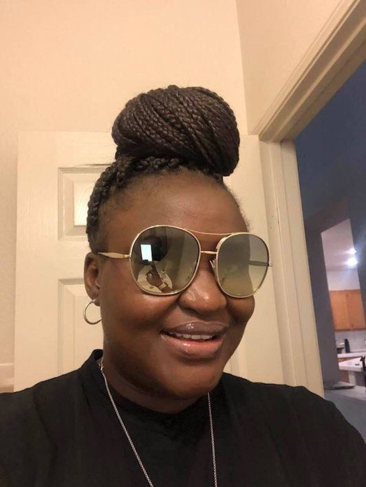 Woman with braided hair in a bun, wearing gold-rimmed sunglasses, smiling. Indoors, against a white door.