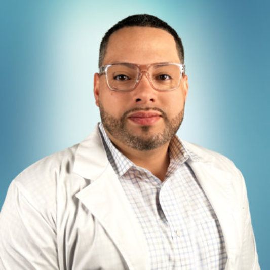 Man in a white lab coat and glasses, smiling, against a blue gradient background.