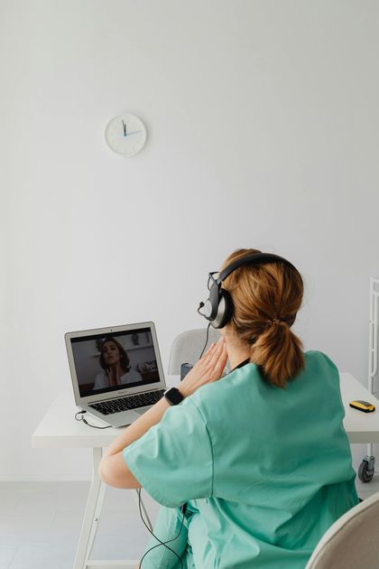 Medical professional in green scrubs, wearing headphones, on a video call with a patient via laptop. White room setting.
