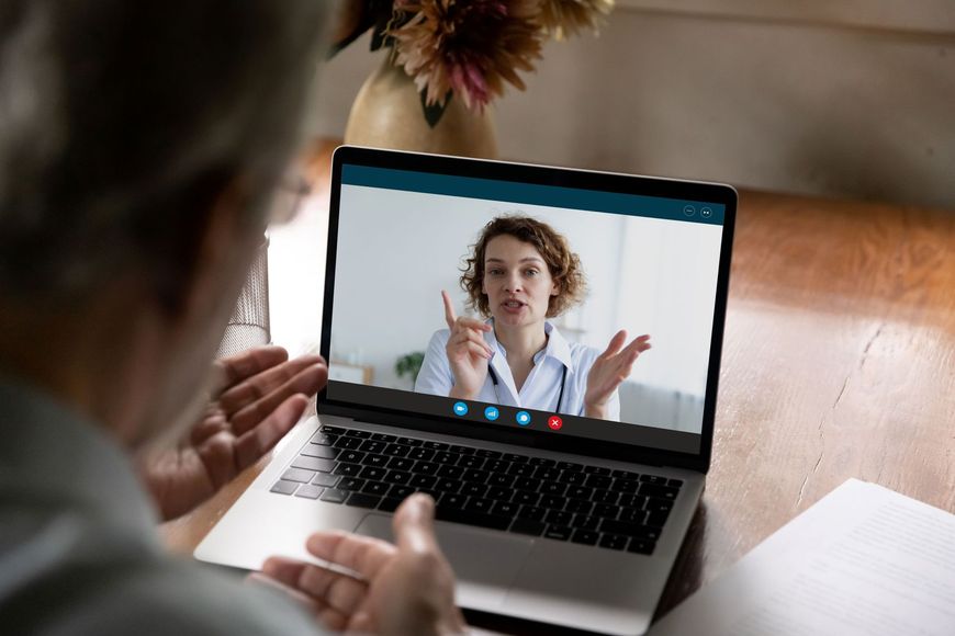 Person on a video call with a doctor on a laptop. The doctor gestures, indoors.