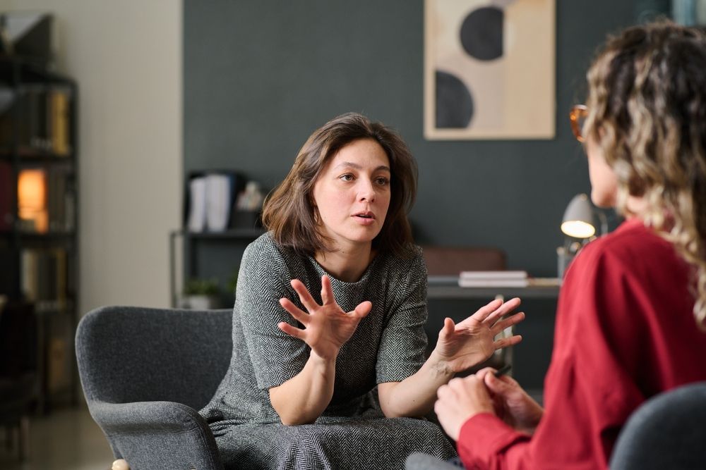 Woman in gray dress gestures, speaking to another woman in a red shirt, in a room; counseling session.
