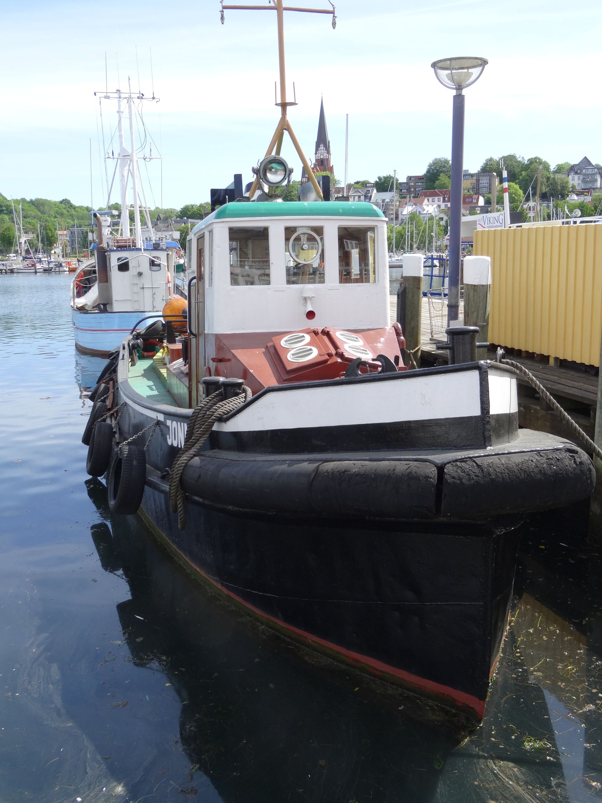 Tugboat docked in harbor, black hull, red stripe, white and red cabin, other boats and buildings in background.