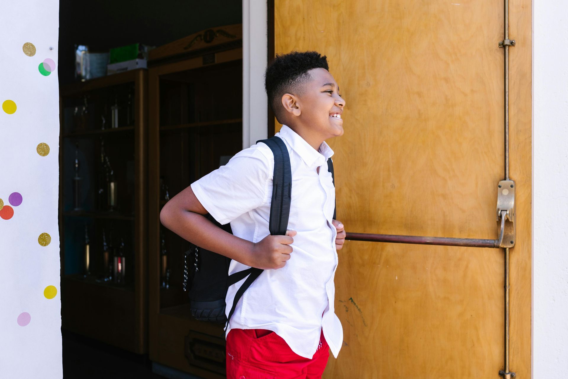 A young boy with a backpack is walking out of a school door.