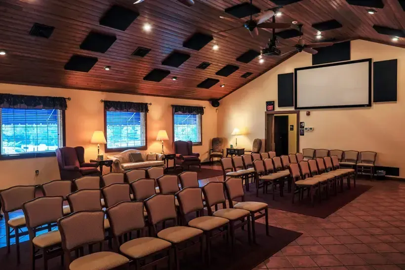Auditorium with rows of chairs, projector screen, windows, and soundproofing panels on the ceiling.