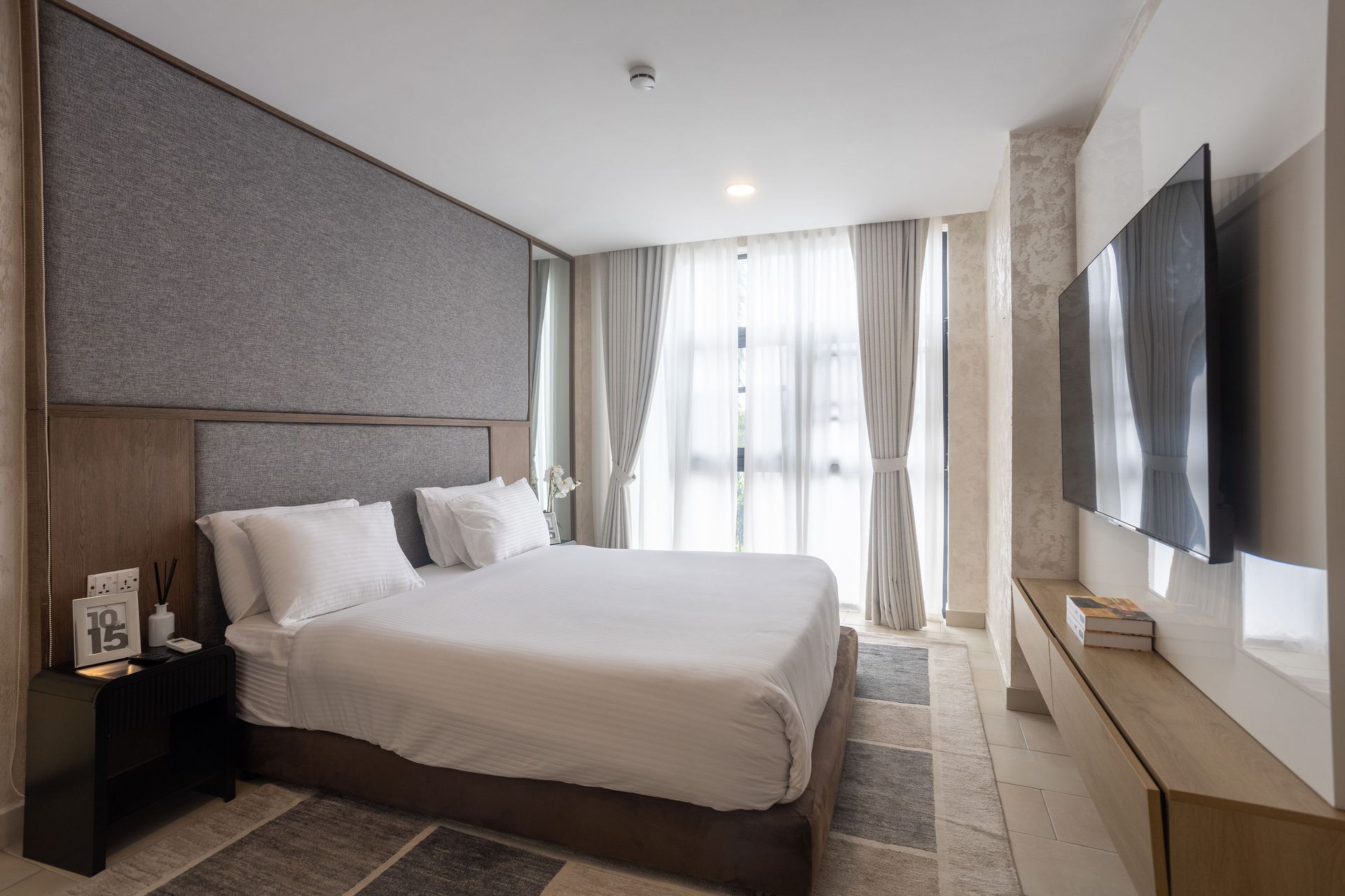 Modern hotel room with a neatly made bed, wall-mounted TV, and bright window with sheer curtains