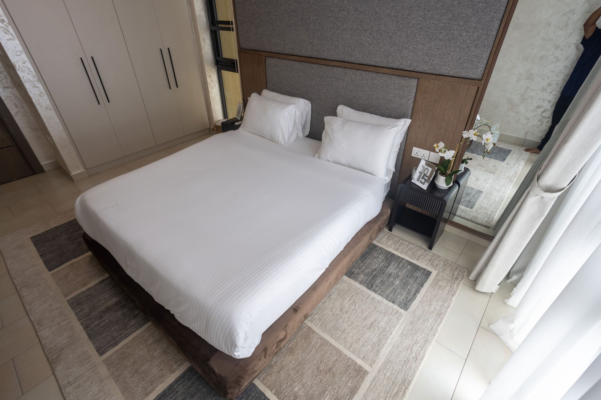 Neatly made bed with white linens in a bright bedroom beside a window and small bedside table