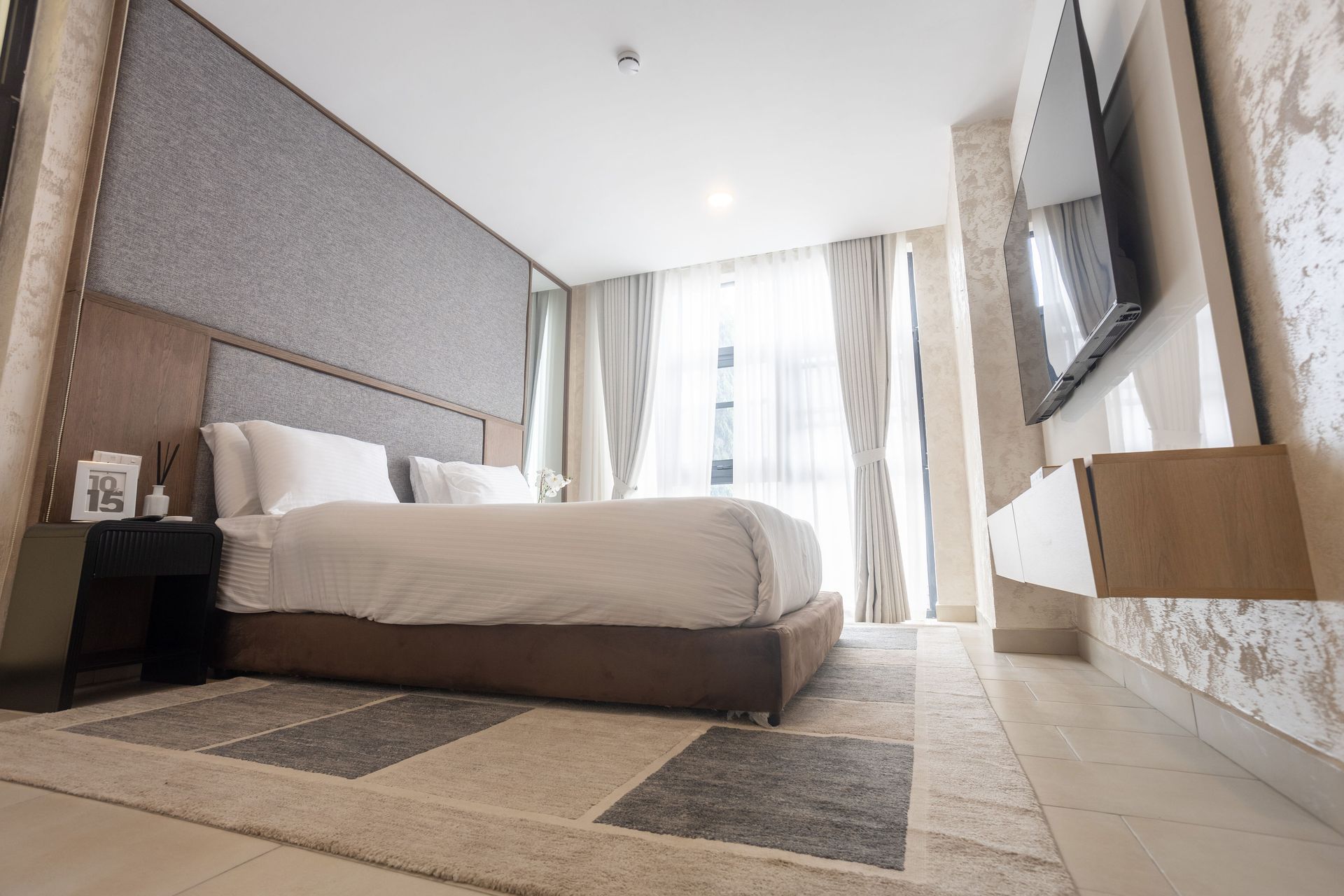 Modern hotel bedroom with a large bed, gray accents, tiled floor, and sheer curtains by the window