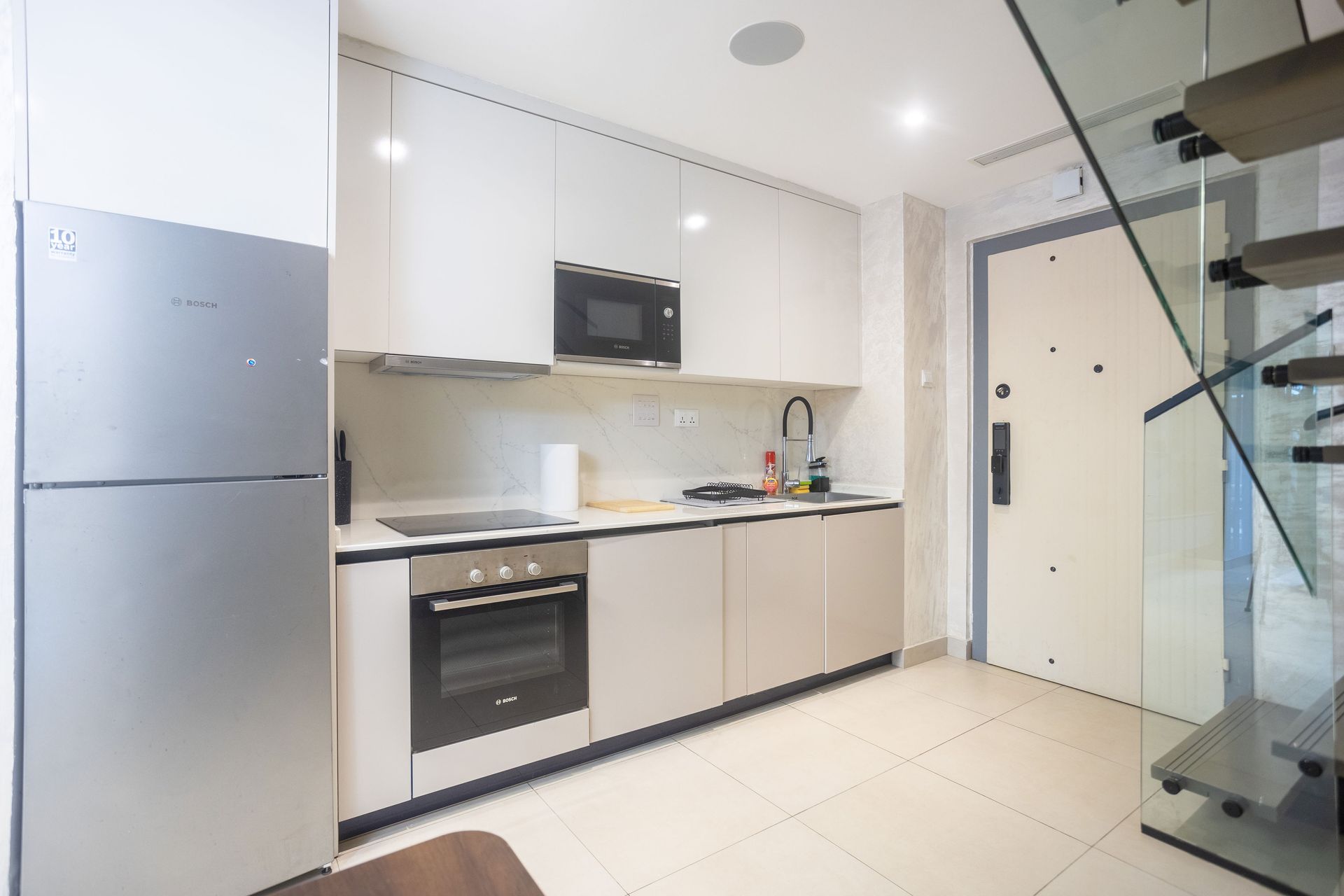 Modern white kitchen with stainless appliances, black countertop, and glass staircase on the right