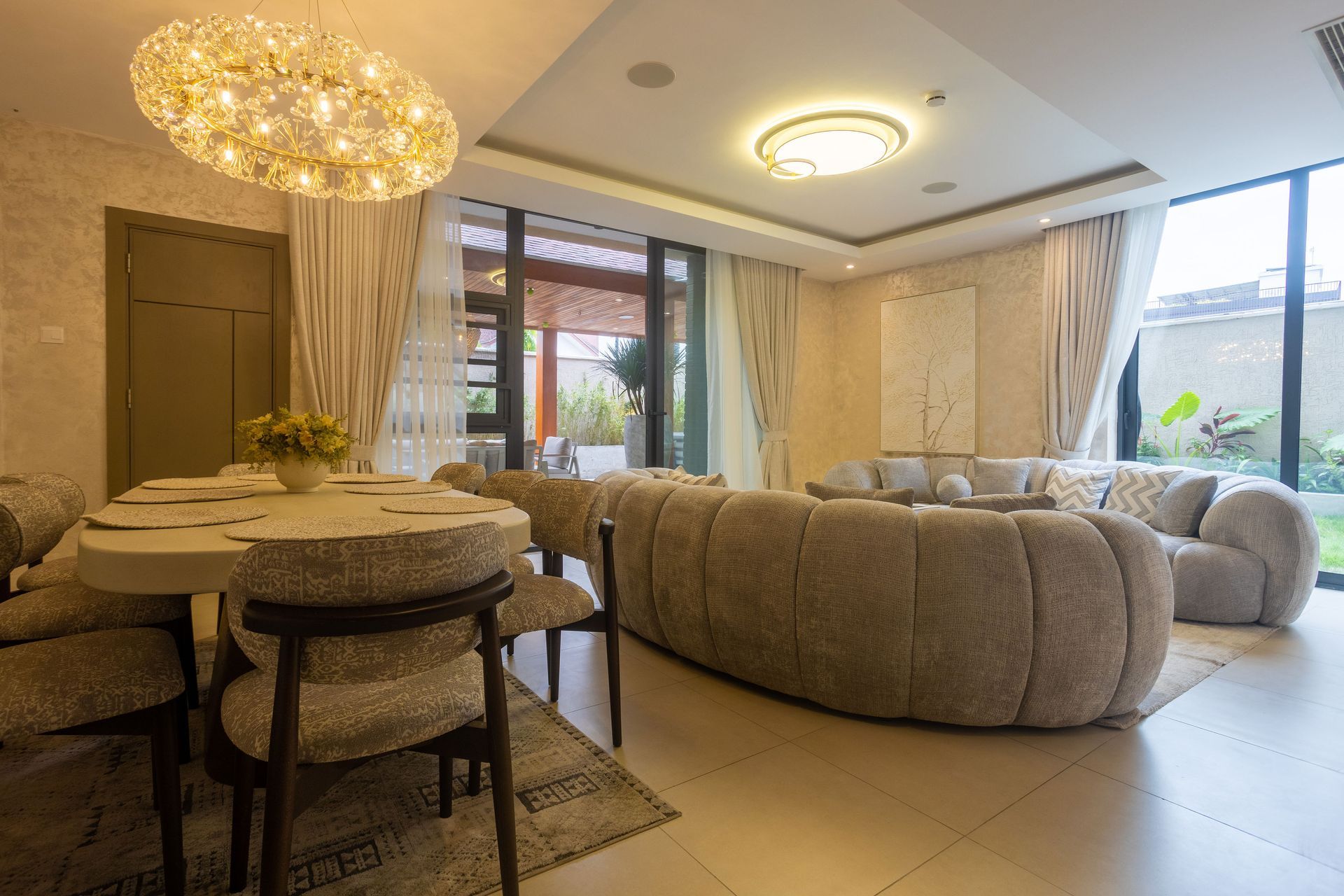 Modern beige living and dining room with curved sofa, round table, chandelier, and large windows